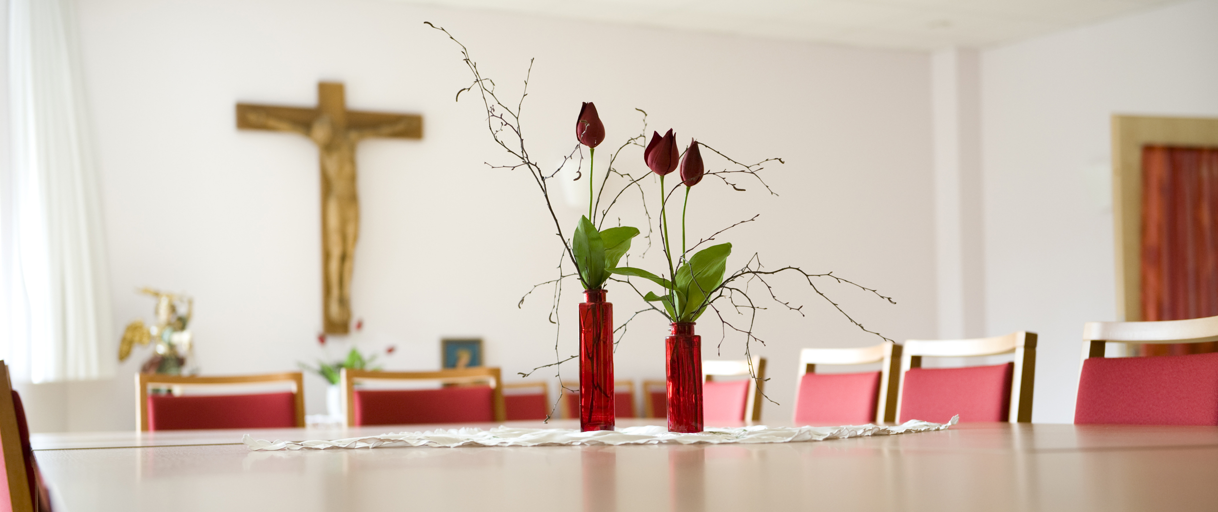 Rosen stehen auf dem Tisch, im Hintergrund hängt ein Kreuz an der Wand.